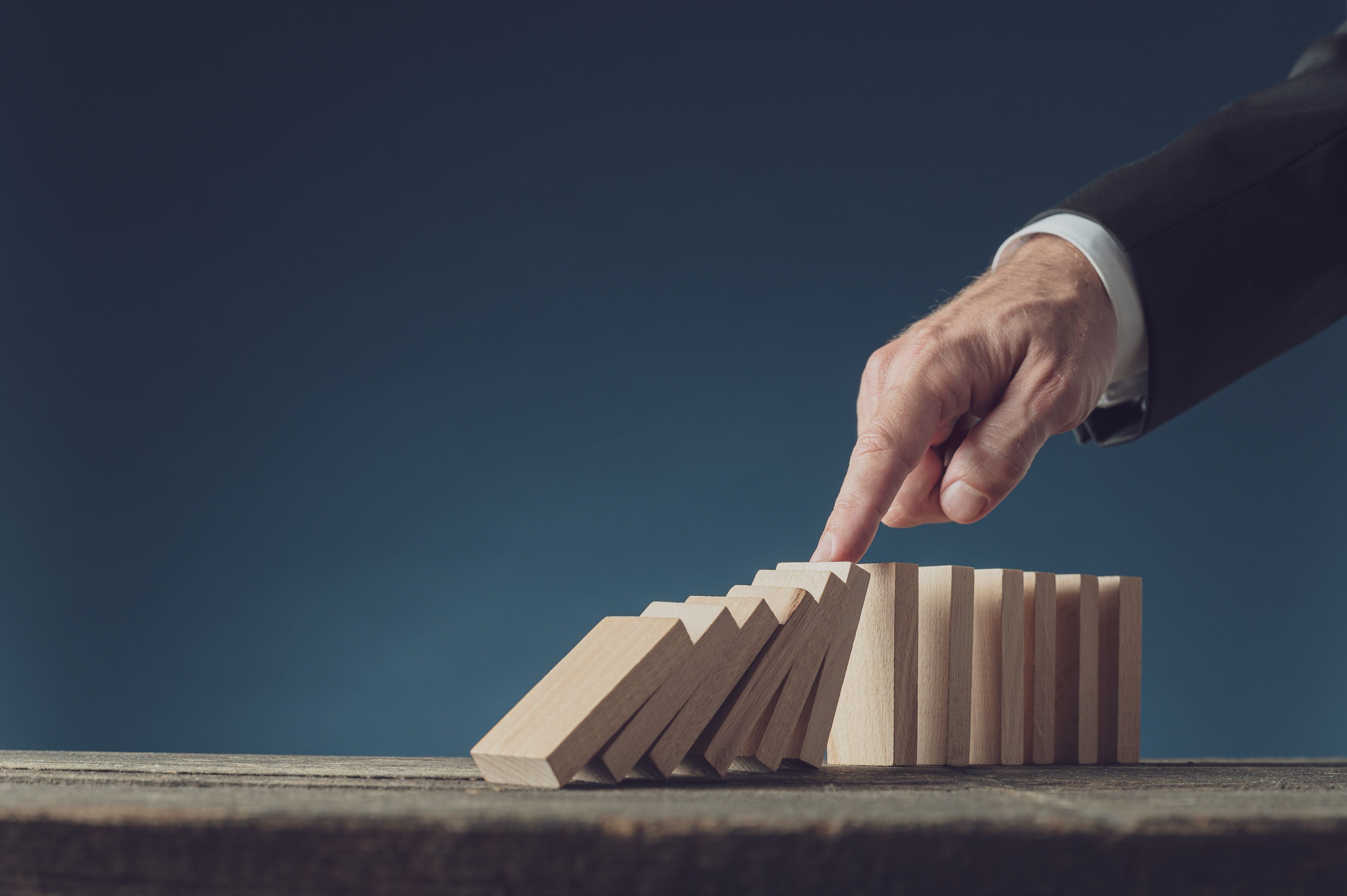 A hand in a suit topples wooden blocks in a domino effect on a wooden table against a dark blue background.